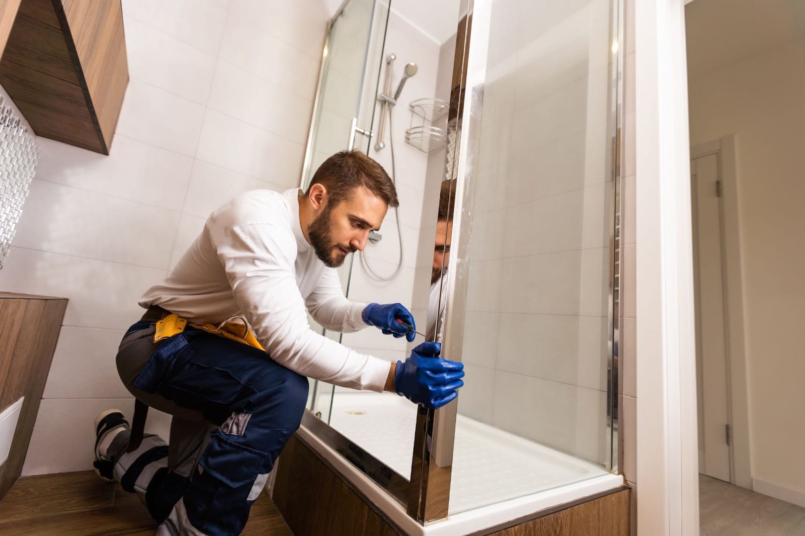 A plumber in a white shirt and blue gloves repairs a shower door, ensuring quality service for your bathtub or shower needs.