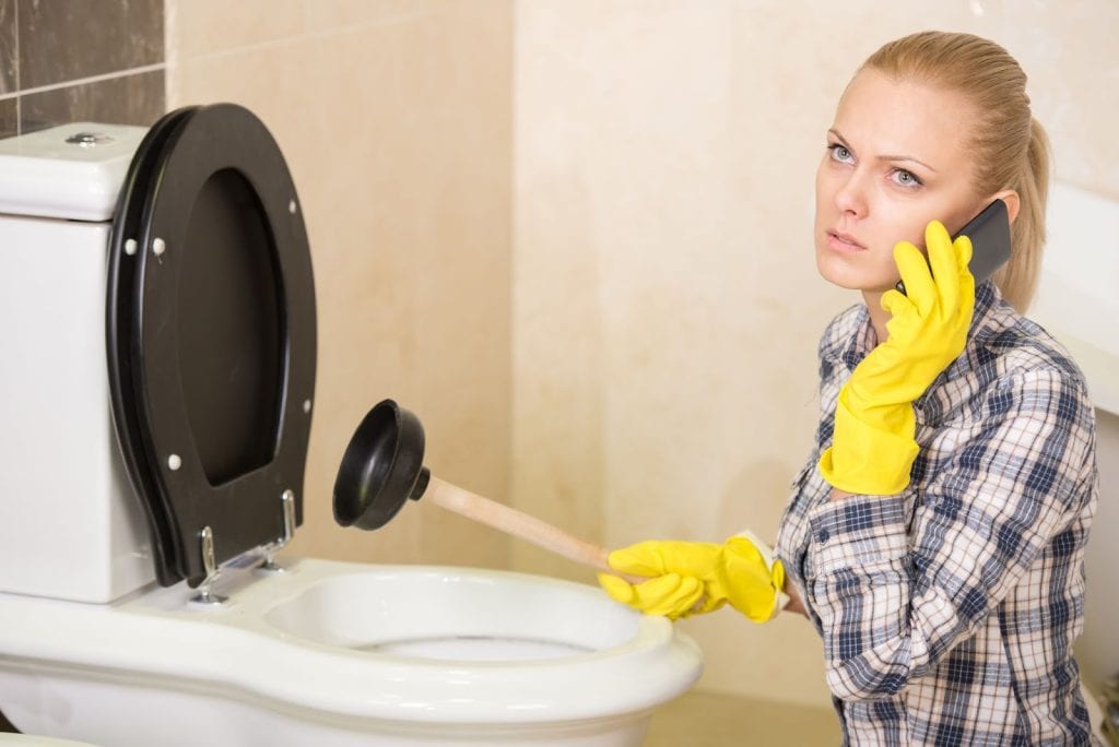 A woman in yellow gloves uses a phone while inspecting a toilet for plumbing repairs in an apartment setting.
