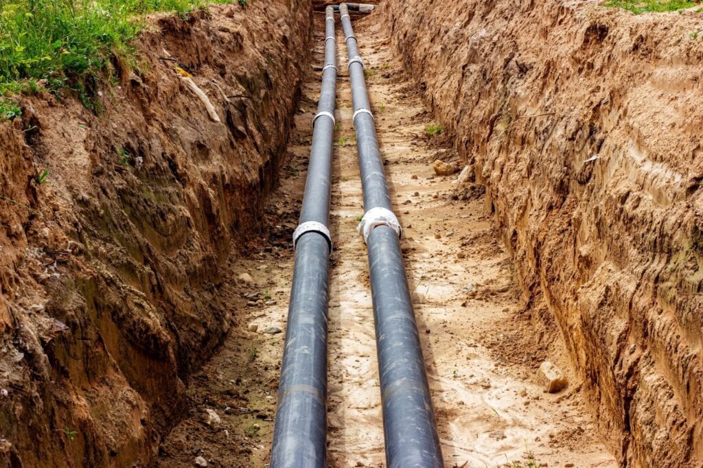 Two parallel black PVC pipes laid in a narrow trench with sandy bedding, secured by white clamps, ready for underground utility installation.