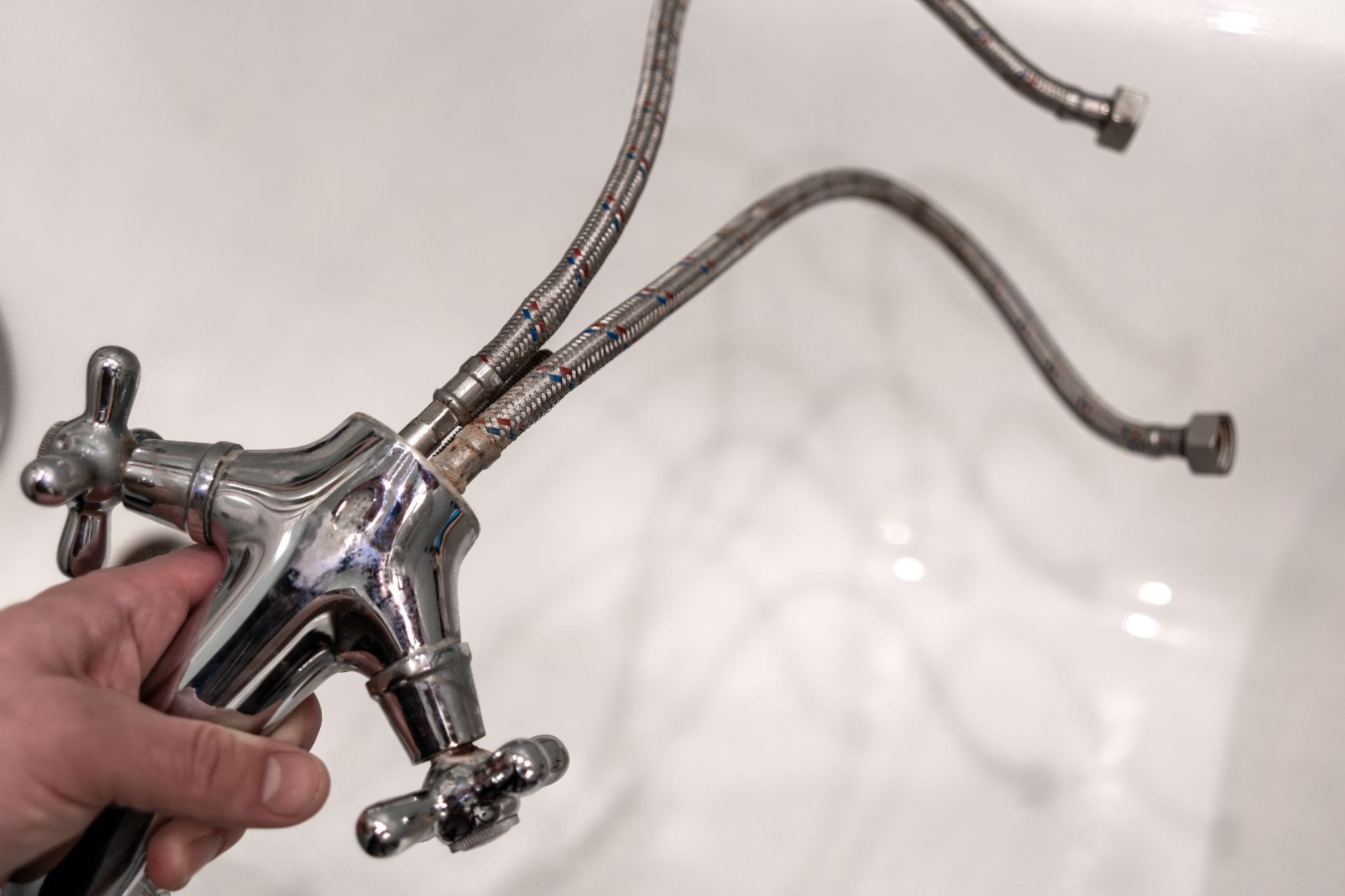 A hand holding a chrome-plated dual-handle faucet attached to long, flexible braided metal water supply hoses.