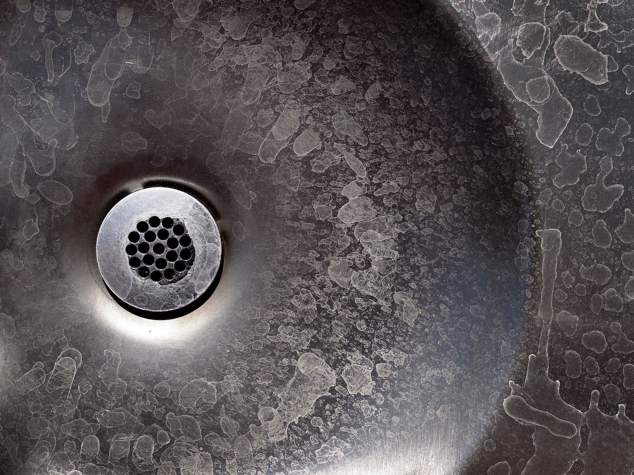 Top-down view of severe white hard water stains and mineral deposits coating a dark metallic sink basin around a drain.