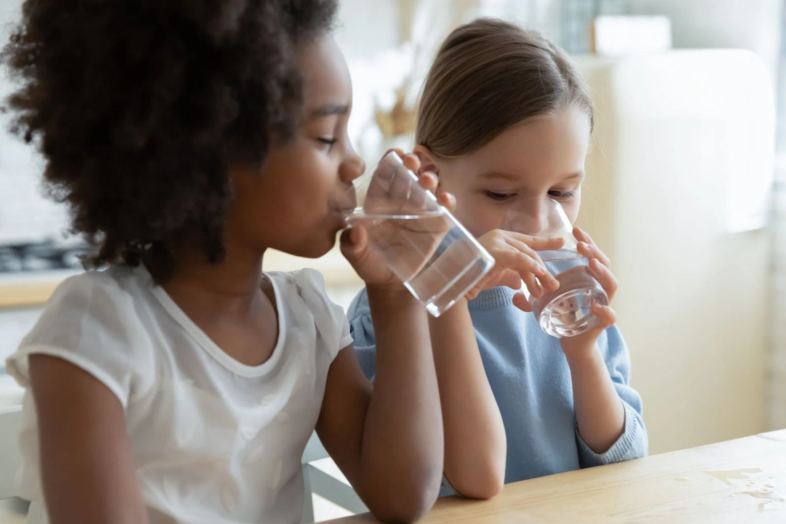 Two children drinking water from clear glasses at a wooden table.