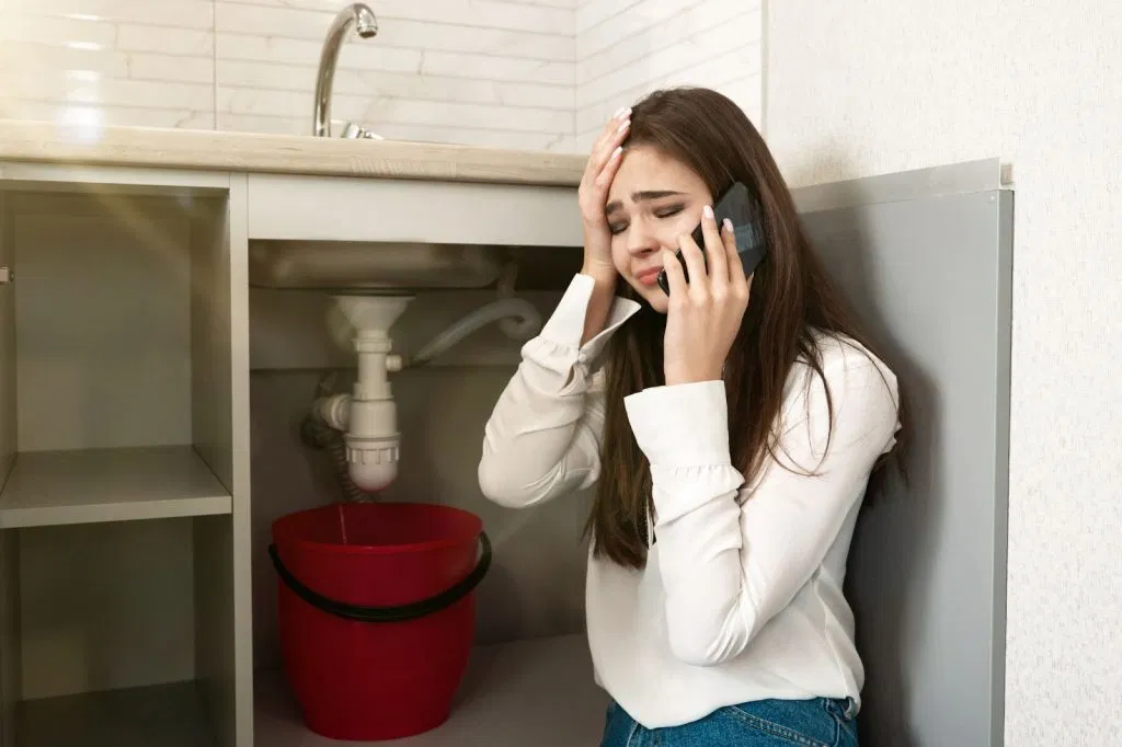 Woman on the phone looking stressed under a kitchen sink with a red bucket nearby.