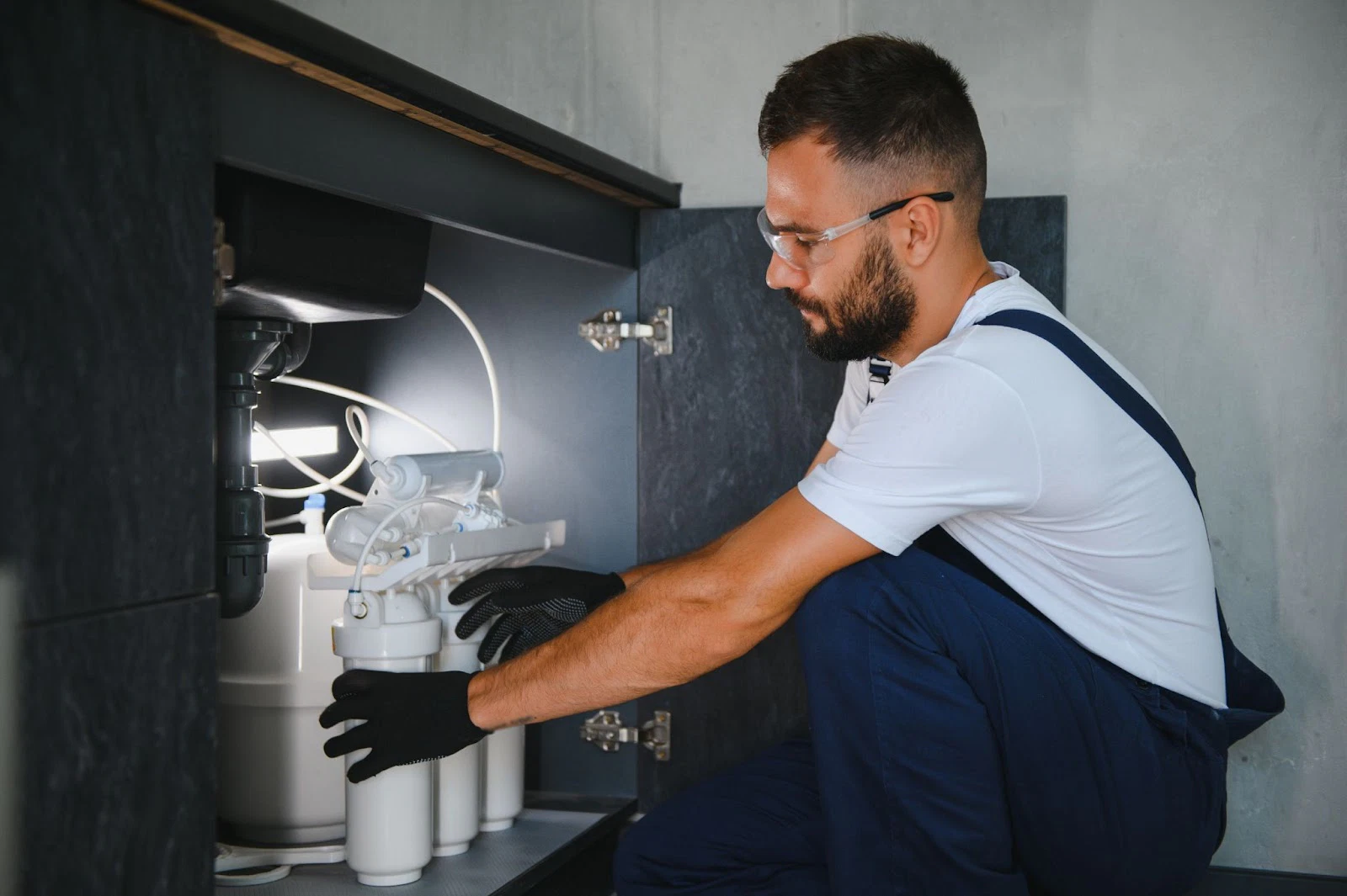 A plumber in overalls and gloves repairing a water filtration system under a sink.