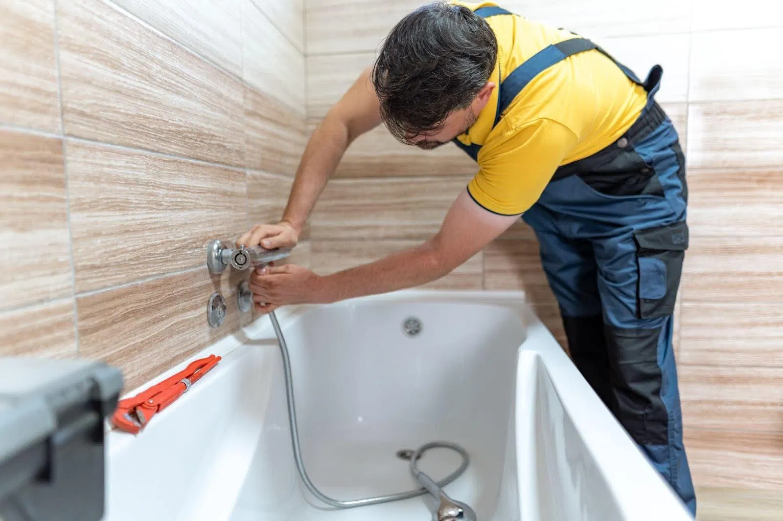A man wearing overalls works on fixing a bathtub in a bathroom, tools visible nearby